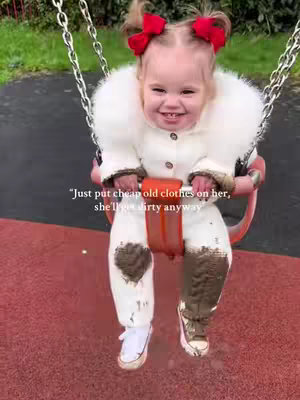 A young girl sits on a pink swing at a playground. She has pigtails with red bows and is wearing a white fuzzy coat and pants. Her pants and shoes are covered in mud. The background shows green grass and trees.