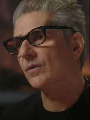 A close-up shot features a light-skinned man with gray hair, wearing black-framed glasses. He is looking to his left with a slightly concerned or thoughtful expression. The image is a medium shot, focusing on his head and shoulders, with the background blurred, suggesting a shallow depth of field.