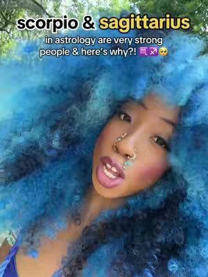 A woman with vibrant blue, voluminous hair is centered in the frame, gazing slightly off-camera with a serene expression. She has multiple facial piercings and is wearing delicate jewelry. The background is blurred, appearing to be outdoors with some foliage visible. The top of the image has overlaid text in white font, and overall the image has soft lighting.
