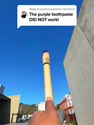 The image shows a wide-angle shot taken from below, looking up at a tall, cylindrical object held by a hand against a clear blue sky. The object resembles a giant tube of toothpaste, featuring a darker purple color at the top end, and a lighter yellow-beige color towards the holding end. A conversation bubble overlaid on the top left corner contains text. In the background, a building and some metallic structures are visible. The composition creates a sense of scale and perspective, with the object appearing large against the sky.