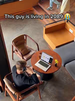 An overhead shot captures a modern interior scene. In the center, a round wooden table hosts a laptop displaying a website, flanked by a small vase of yellow flowers. A man sits at the table, facing away from the camera. To the left, a brown wooden chair with a leather seat is visible. In the background, a couple is walking up the stairs. The overall setting is a cafe or working space designed in a modern style with brown tones and a gray couch.