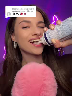 A woman with long brown hair leans forward, applying foamy substance from a white and blue can directly into her open mouth. She is wearing a dark top and large pearl earrings.