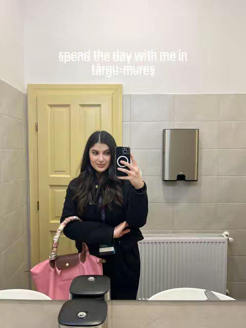A woman takes a selfie in a public restroom, holding her phone up to the mirror. The background includes a beige tiled wall, a gray metal hand towel dispenser, a white porcelain sink and a closed door, and a radiator. She holds a pink tote bag. Text overlay "spend the day with me in targu-mures" is at the top of the image.