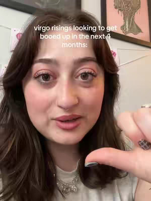 A selfie-style video shows a woman with dark hair and makeup, looking directly at the camera. Text overlays near the top of the screen announce a support group. The background is lightly decorated with art. The woman is wearing a necklace and making a gesture with her hand.