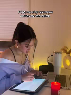 A person sits at a desk, illuminated by soft lighting. A large tablet with a lit screen is in front of them. A candle burns to the side, and a laptop is also present. The person has dark hair pulled back in a ponytail, and a light-colored button-down shirt.