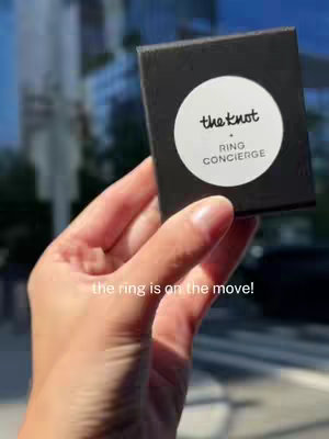 A hand holding a black box with a white circular sticker. The sticker has the words "the knot" in a handwritten font, and the words "RING CONCIERGE". The background is blurred, showing a cityscape.
