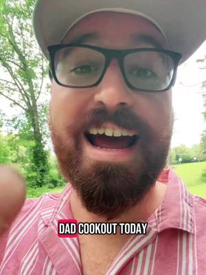 A man with a beard, wearing glasses, a hat, and a button-down shirt, smiles widely at the camera. Text overlayed at the bottom of the image reads 'DAD COOKOUT TODAY'. The background is blurred with green trees and grass.