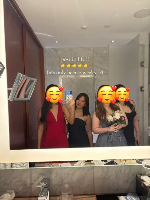 A mirror selfie shows four young women posing in what appears to be a hotel room, likely taken during a formal occasion. The women are framed in the mirror, each wearing elegant dresses. The room is well-lit, with a modern design, and a bathroom area is visible in the background. A text overlay at the top of the image reads, 'post ib life !!' with several emojis indicating the end of their student days. The overall composition gives a sense of celebration and the end of an academic period with those who seemed to experience the shared journey.