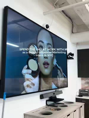 The image is a medium shot of a large flat-screen television mounted on a white wall. The screen displays a video featuring a woman demonstrating putting on makeup. The text overlay in the image reads, 'SPEND THE DAY AT WORK WITH ME as a Beauty Influencer Marketing Intern In NYC.' Below the screen is a counter-height cabinet with built-in trash receptacles. Two black security cameras are mounted near the top of the screen, and a black speaker/microphone setup is installed below the TV.