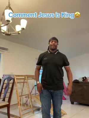 A man stands in what appears to be a living room, dressed in a black polo shirt and jeans. Above him, text reads, "Comment Jesus is King" in a bubble. A chandelier and furniture are visible in the background.