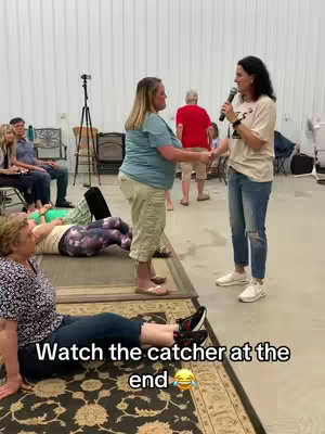 The image shows a group of people indoors, possibly in a large room or hall. A woman with a microphone is standing, addressing the others while another woman is standing near her. The scene appears to be an interactive event or gathering, likely a group session or presentation. A rug with a pattern is located in the lower center. There are people in the background as well.