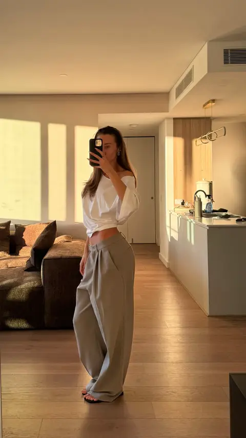 A woman in a white off-the-shoulder top and wide-leg gray pants is taking a mirror selfie. She is holding her phone up to her face, partially obscuring it, and the background shows a modern apartment with a kitchen, a sofa, and sunlight streaming in.