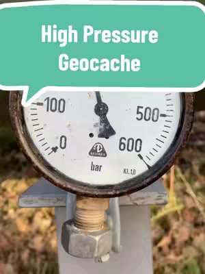 The image shows a close-up of a pressure gauge on a metal post. The gauge is round with a white face, black numerals, and a black needle pointing near the 600 mark. The gauge face has the text "bar" and "KL.10". A blue speech bubble with the text "High Pressure Geocache" floats above the gauge.