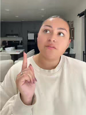A woman in a light-colored sweater points her index finger upward with a thoughtful expression. The background suggests a home setting with a kitchen and living area visible.
