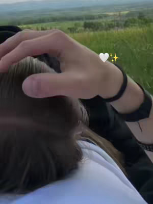An overhead view of a couple outdoors. The primary focus is a hand softly touching the head of someone, presumably the other person. In the background is a green grassy area, and trees. There is also an emoji heart with sparkle effects positioned beside the hand.