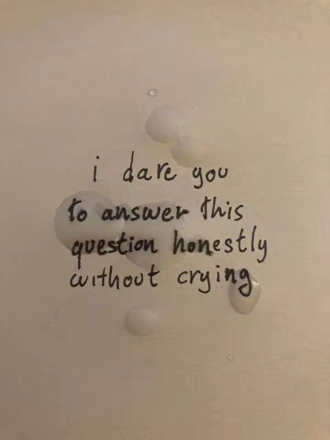 A close-up shot of a white surface with handwritten text in black ink, and some water droplets or stains. The text reads: 'I dare you to answer this question honestly without crying.' The composition is simple, with the text centered and the droplets scattered around it.
