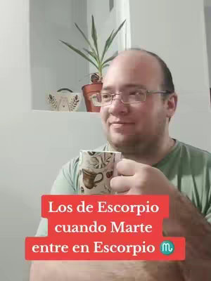 A man, wearing glasses and a green t-shirt, is holding a coffee mug, looking sideways with a gentle smile. Behind him is a cabinet with a plant on top.