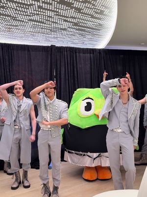 The image features multiple young men in matching light gray military-style outfits, posing with a large, green Duolingo mascot. They are in a well-lit indoor space with a bright circular patterned ceiling feature and black curtains in the background. The men are performing a dance pose, with one arm raised and hand on their head. The Duolingo mascot is in the center and the entire combination creates a promotional feel for the language learning app.