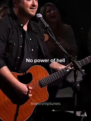 A man playing an acoustic guitar and singing into a microphone. The image has a dark, theatrical lighting with a blurred background featuring another person. Text overlayed at the bottom displays the account name.