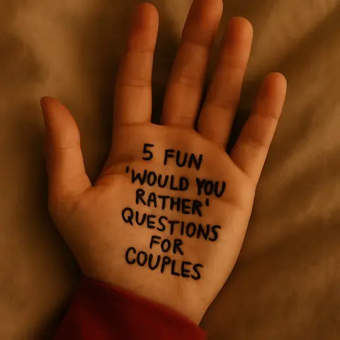 A close-up of a hand, palm facing up, with text written on it. The hand is fair-skinned and resting on a textured, light brown surface. Black ink is used to write: "5 FUN 'WOULD YOU RATHER' QUESTIONS FOR COUPLES." The angle is slightly from above, emphasizing the text on the palm.