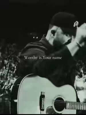 A black and white image featuring a man playing an acoustic guitar with a crowd in the background, a text overlay says 'Worthy is Your name'.