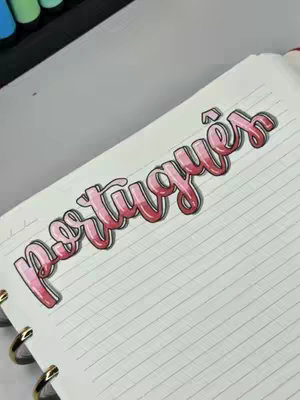 A close-up shot of a notebook page with the word "Portugues" elegantly written in pink brush lettering, accented with a black outline. The word is centered on the lined paper, with colorful markers visible in the upper left corner.
