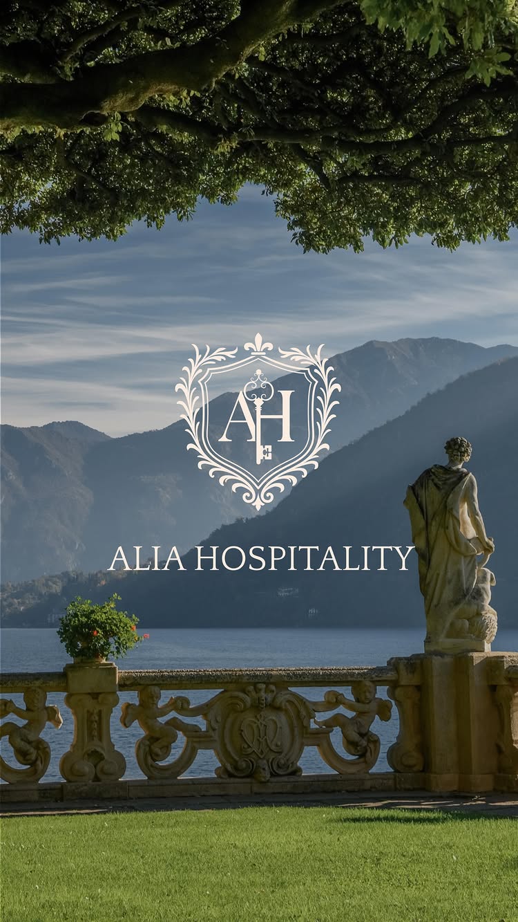 The cover image showcases a serene outdoor scene dominated by a tranquil lake and distant mountains, framed by a lush green foreground. In the foreground, a stone balustrade with ornate carvings and sculptures leads the eye. A classic statue faces the lake, enhancing the elegance. The sky is clear, hinting at a beautiful day. The logo 'ALIA HOSPITALITY', in elegant font, is positioned at the bottom. Overall, the image evokes a sense of luxury, tranquility, and scenic beauty.