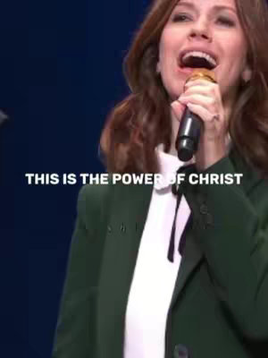 The image shows a woman singing into a microphone on stage, with text overlayed on the foreground. She has reddish-brown hair and is wearing a white collared shirt and green blazer. The background is a dark blue color. The text overlay says "THIS IS THE POWER OF CHRIST".