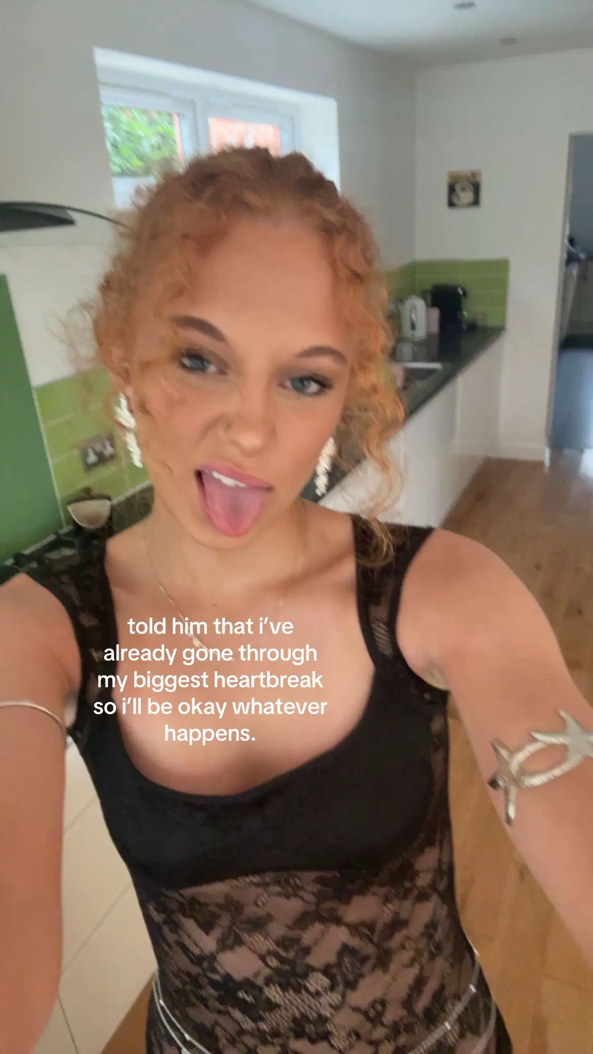 A selfie-style shot with a young woman with curly red hair and blue eyes as the focal point. She is wearing a black top with lace detailing, and the background shows a kitchen setting with neutral-colored walls and some green accents. Text overlays the image, conveying a message about relationship resilience.