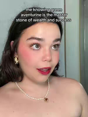 A close-up shot of a woman with fair skin, dark hair, and red lipstick. She is wearing a pearl necklace, gold earrings, and her face is flushed with bright pink blush. Text overlayed on the image says, "me knowing green aventurine is the master stone of wealth and success."