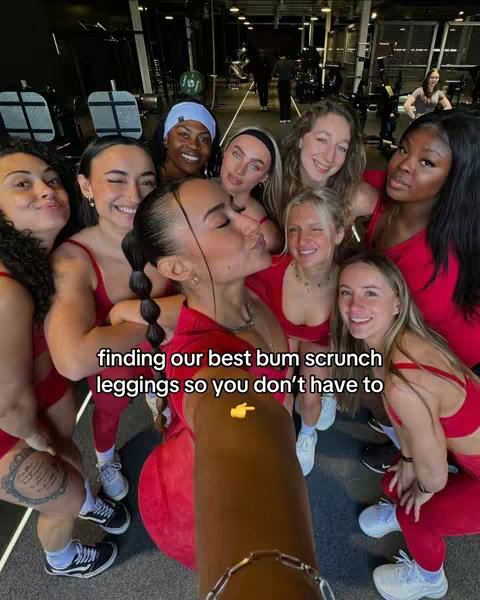 A group selfie in a gym setting. Around ten women are gathered, predominantly in red workout attire. The central figure is closest to the camera, surrounded by others with varying skin tones and hairstyles. The overall composition is vibrant, with gym equipment subtly in the background.