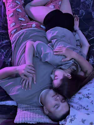 Two people, likely a couple, are embracing while lying down, with their heads close together. They are in bed, with a floral design pillow and other bedding visible. The man at the bottom has his eyes closed, while the woman at the top is looking down. The lighting has a purple hue.