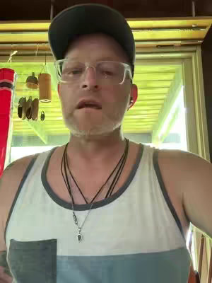A man in a tank top and cap is looking at the camera. He is wearing glasses and a necklace.