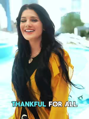 A woman with long black hair and a gold necklace smiles widely, set against a blurred, light blue and green background that suggests a pool or outdoor setting. She wears a yellow, sheer top over a black garment, and a turquoise text overlay at the bottom reads 'THANKFUL FOR ALL'.