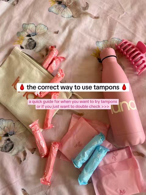 A top-down shot shows a collection of menstruation products arranged on a patterned pink floral fabric. The items include a pink water bottle, a hair clip, pouches, and individually wrapped tampons. Text highlights the topic of the video.