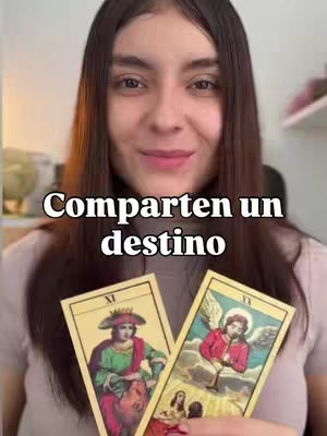 A woman with long brown hair smiles slightly, holding two tarot cards in front of her. Above her, the text 'Comparten un destino' (They share a destiny) floats in a white, bold font. The cards depict figures with elaborate clothing, in shades of beige, green, red, and gold against a soft, blurred background, focusing the attention on the cards and her face.