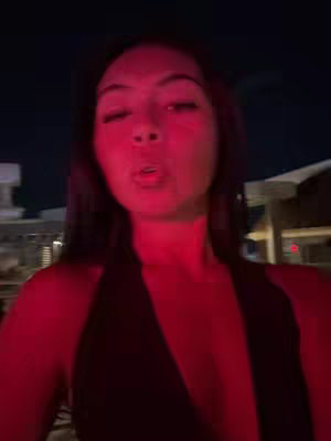 A selfie taken at night with a woman positioned in the center. The lighting is predominantly red, casting a strong hue over her face and the surrounding environment. She appears to be outdoors, with indistinct structures in the background. Her expression is somewhat playful, possibly blowing a kiss.