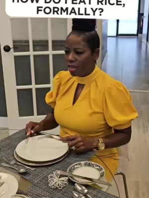 A woman in a yellow dress is seated at a table set for a formal meal. She has dark hair pulled up and is looking toward the left of the frame. A table setting with plates, silverware, and a small decorative element are visible.