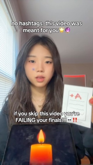 A young woman with dark hair and fair skin is centered in the frame. She looks directly at the camera with a slightly concerned expression. The image is composed with text overlays at the top and bottom, framing her face. Behind her is a blurred interior setting. She holds a flashcard with an 'A' grade. Below, a candle projects its flame onto a tablet surface.