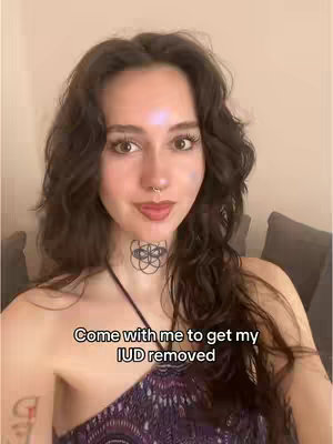 A woman with long, wavy dark hair smiles at the camera. She is wearing a low-cut top and has a tattoo on her neck. Text overlayed reads: "Come with me to get my IUD removed."