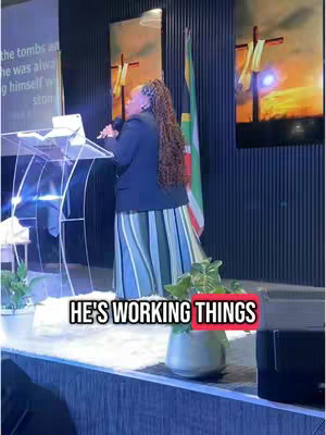 A woman in a black blazer and long skirt is speaking from behind a clear podium in front of a dark gray wall. The hook text is in a bright red box. Green plants in a silver pot in front of the stage.
