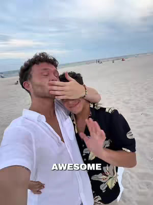 Two young men pose on a beach. One covers the other's eyes with a hand, and the other seems to be waving with his other hand. Text is displayed in large, white font across the bottom of the image.