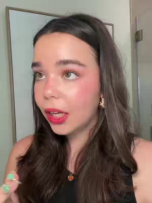 A young woman with light skin and brown hair is speaking to the camera while wearing a black tank top and a small pendant necklace. Her makeup includes pink blush and red lipstick. She has a green stone ring on her finger and earrings in her ears. Her non-dominant hand is holding a green ring.