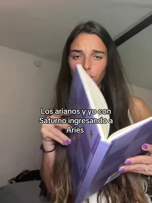 A woman with long dark hair is seen holding a purple book open, covering part of her face. She is looking down with a thoughtful expression. Text overlay is visible at the bottom left that reads: "Los arianos y yo con Saturno ingresando a Aries". The setting appears to be indoors, with a muted color palette.