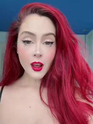 A woman with bright red hair and dramatic makeup looks towards the camera with an open mouth, her eyes wide. The background is a solid blue, and she has a black top on. The makeup includes winged eyeliner and the lip color is a bright red.