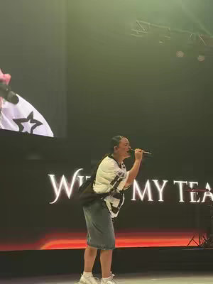 A person with short, dark hair, wearing a white short-sleeved shirt over a black and white vest, and a denim skirt, sings into a microphone onstage. The stage has a dark red screen with text on it, and lighting effects are on from above and to the side. A partial flag can be seen in frame, black outline of a star is seen on the flag.