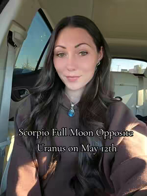A woman with long, wavy dark hair is seated in a car, facing the camera with a gentle smile. She wears a brown hoodie and a necklace with a blue pendant. The text overlay reads, 'Scorpio Full Moon Opposite Uranus on May 12th.' The background suggests an outdoor setting with natural light coming through the car window.