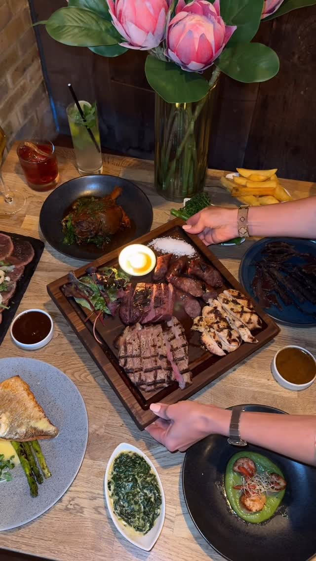 A wooden table is set with multiple plates of food, showcasing a variety of culinary delights. The main focus is a wooden tray holding grilled meats, including steaks, sausages, and chicken, along with a lit candle. Side dishes and saucers are scattered around the tray, including a plate of fried potatoes, greens, and dipping sauces. Floral arrangement and drinks are visible as well.