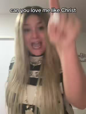 A woman, with long blonde hair and a black and white top, is in the foreground with her face centered. She has an open mouth expression and is gesturing with her right hand, showing a middle finger. The background is out of focus, with a white wall and a glimpse of an open doorway to the left.