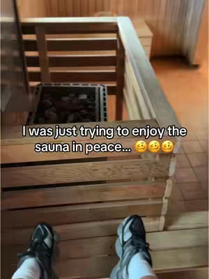 The image shows a low-angle view inside a sauna. The foreground features the feet of a person wearing black and white sneakers, positioned on wooden steps. The middle ground shows sauna benches and the sauna stove. The background showcases the sauna's wooden structure, creating a warm, rustic atmosphere. The overall scene is bathed in warm, inviting tones from the sauna lighting.